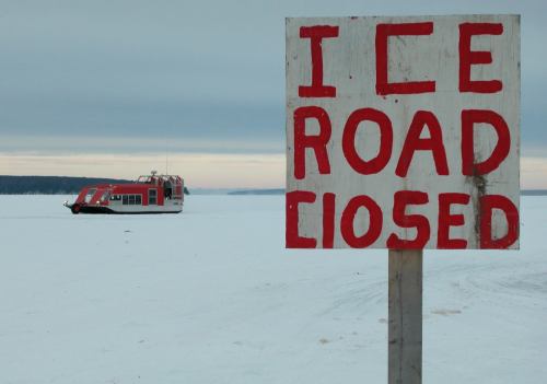 A sign reading "ICE ROAD CLOSED" stands in front of a red vehicle on a snowy, frozen landscape.