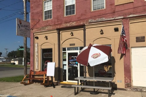 Exterior of a BBQ restaurant with an open sign, picnic table, and an umbrella, featuring a nearby American flag.