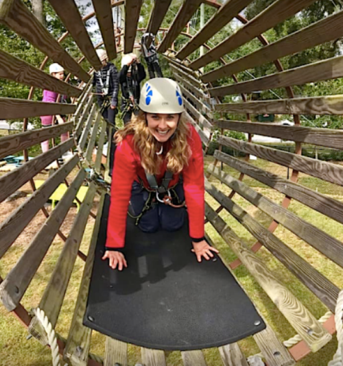 A woman in a helmet smiles while crawling through a wooden tunnel at an outdoor adventure park.