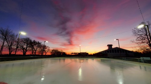 A serene ice rink at sunset, with vibrant colors in the sky and trees silhouetted against the horizon.