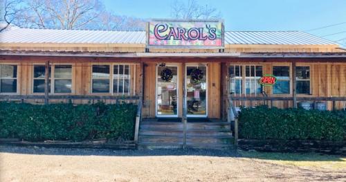 A wooden storefront with a sign reading "Carol's" and an "Open" neon sign, surrounded by greenery.