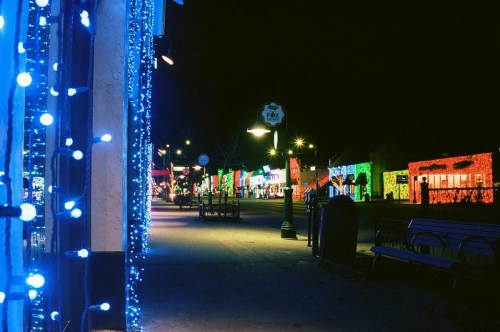 A festive street at night, adorned with colorful holiday lights and decorations along the buildings and trees.