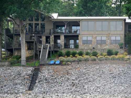 A large stone and wood house by a lake, featuring a deck, stairs, and landscaped gardens along a rocky shoreline.