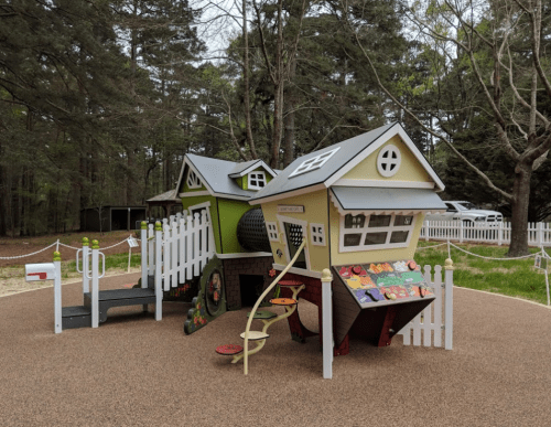 Colorful playground structure resembling two houses connected by a slide, surrounded by trees and a white picket fence.