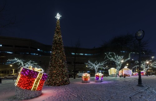 A festive scene featuring a lit Christmas tree and large, colorful gift boxes in a snowy park at night.