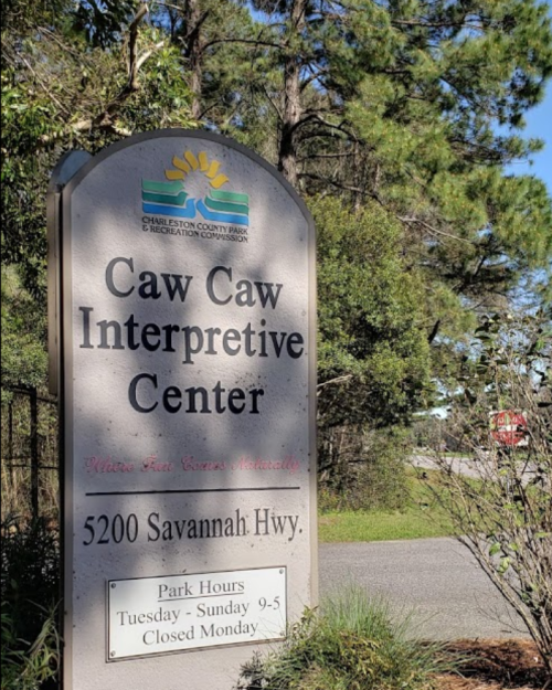 Sign for Caw Caw Interpretive Center, listing hours and location at 5200 Savannah Hwy, surrounded by trees.