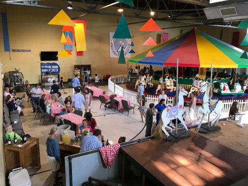 A lively indoor event with people socializing, a carousel, and colorful decorations in a festive atmosphere.