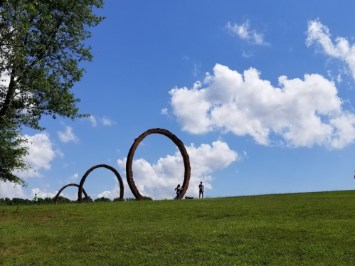 Three large circular sculptures on a grassy hill under a blue sky with fluffy clouds, with people in the distance.