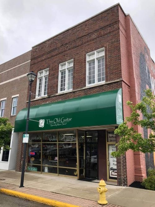 Storefront of "This Old Guitar" with green awning, showcasing musical instruments and a brick building exterior.