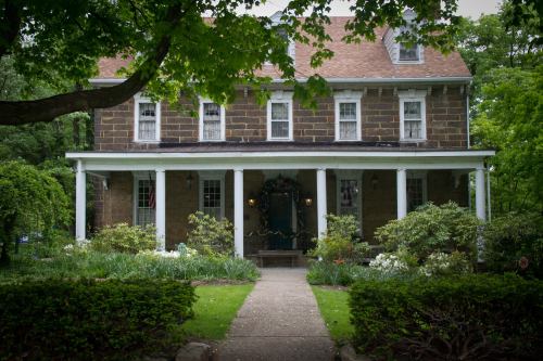 A charming two-story stone house with a front porch, surrounded by lush greenery and colorful flowers.