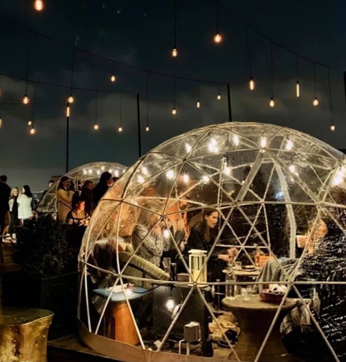 A cozy outdoor setting with illuminated igloo-like structures, people enjoying drinks under string lights at night.