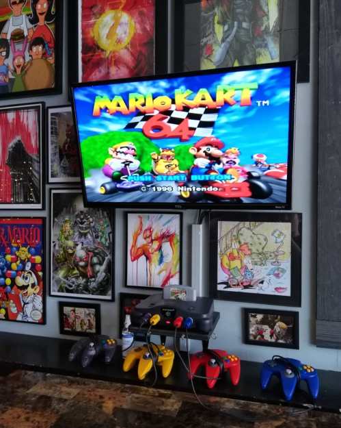 A TV displaying the title screen of Mario Kart 64, with colorful game controllers and framed artwork on the wall.