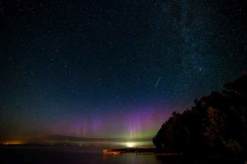 A starry night sky with vibrant auroras and a calm lake, framed by trees and a distant pier.