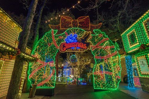 Colorful holiday lights form a festive archway with "Christmas Midtown" sign, surrounded by decorated buildings at night.