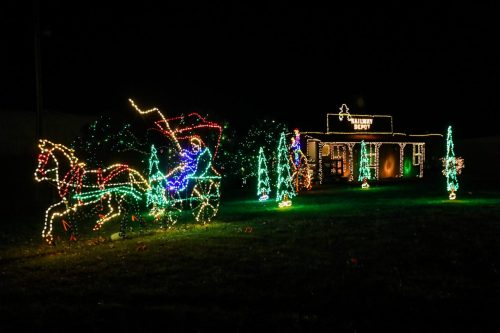 Colorful holiday lights depict a horse-drawn carriage and a festive building surrounded by illuminated trees.