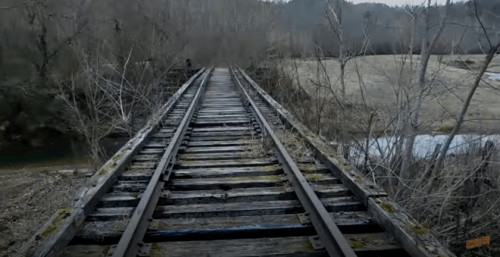 A weathered wooden railway bridge stretches over a river, surrounded by trees and a grassy landscape.