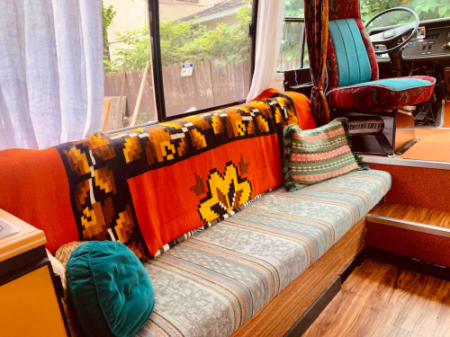 Cozy interior of a bus with colorful cushions, a patterned blanket, and a driver's seat visible in the background.