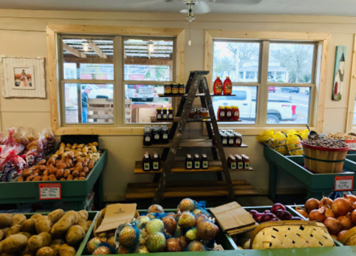 A cozy market interior with fresh produce, jars of honey, and a ladder, featuring large windows and a view outside.
