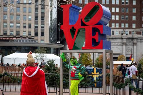 A festive scene featuring a large "LOVE" sculpture with a person dressed as the Grinch in a holiday sweater.