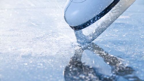Close-up of a skate blade gliding over ice, creating splashes of water and reflections on the surface.
