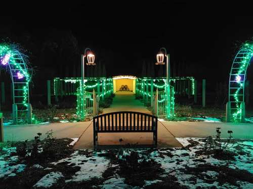 A pathway lined with green lights and arches, leading to a lit entrance, with a bench in the foreground at night.