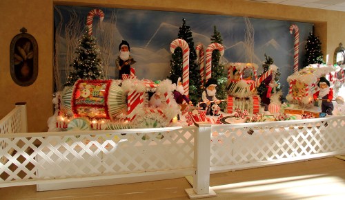 A festive display featuring a train made of candy, decorated trees, and holiday figures against a snowy backdrop.