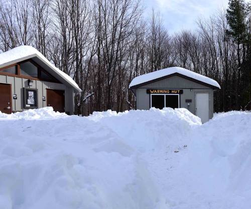 Two snow-covered huts surrounded by deep snow, with a sign reading "Warming Hut" on one of the buildings.