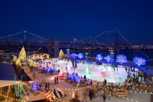A festive ice skating rink at night, surrounded by twinkling lights and decorated trees, with a bridge in the background.