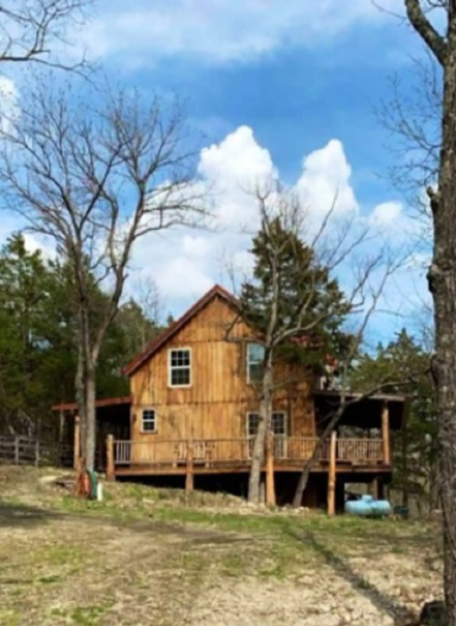 A rustic wooden house surrounded by trees under a blue sky with fluffy clouds.