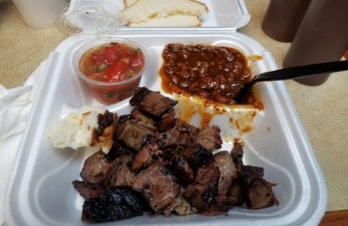 A takeout container with smoked meat, baked beans, salsa, and a piece of bread on a table.