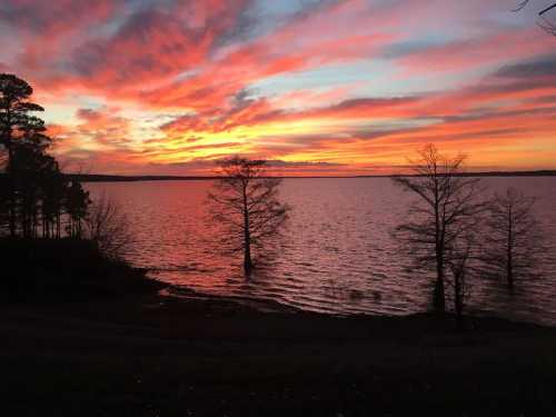 A vibrant sunset over a lake, with colorful clouds and silhouetted trees along the shoreline.