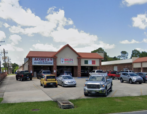 A storefront with "Acosta's" and "Rocco's" signs, surrounded by parked cars and a blue sky with clouds.