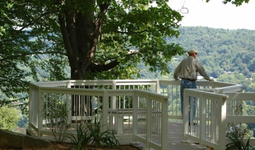 A person stands on a wooden deck overlooking a scenic valley, surrounded by trees and greenery.