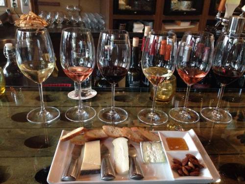 A selection of wine glasses and a cheese platter with bread, nuts, and honey on a bar counter.