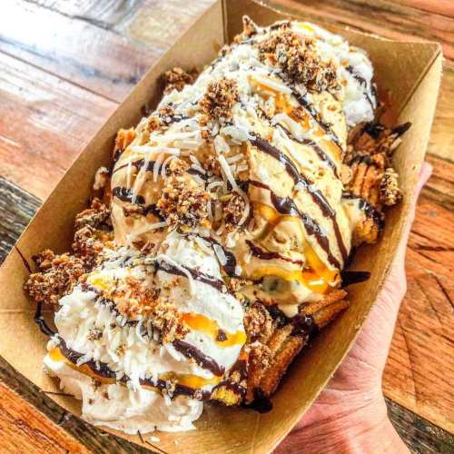 A delicious dessert featuring ice cream, whipped cream, chocolate drizzle, and crunchy toppings in a tray.