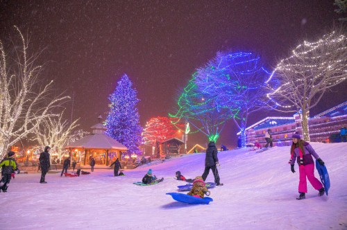 A snowy scene with colorful lights, people sledding, and a decorated tree in a festive winter atmosphere.