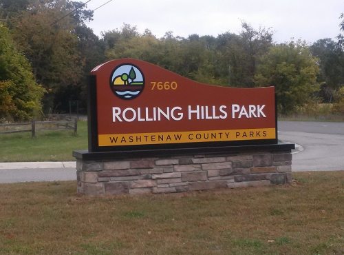 Sign for Rolling Hills Park, Washtenaw County Parks, featuring a logo and the address 7660. Greenery in the background.