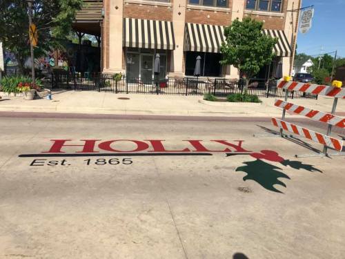 Street view featuring "HOLLY Est. 1865" painted on the pavement, with construction barriers and nearby buildings.