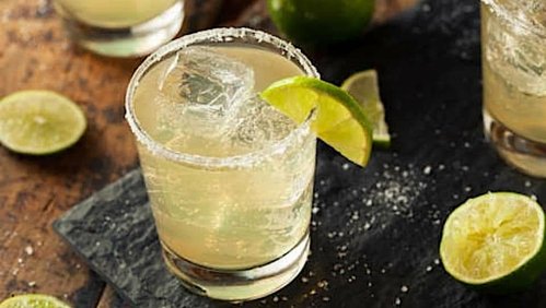 A refreshing margarita in a glass with ice and a lime wedge, surrounded by lime halves on a dark slate surface.