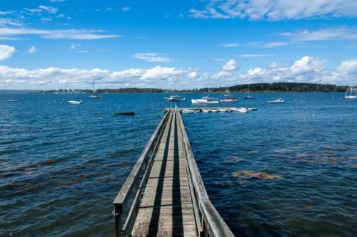 A wooden pier extends into a calm blue sea, with boats scattered in the water under a bright, partly cloudy sky.