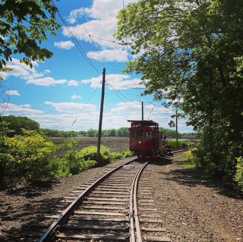 A red train on a winding track surrounded by greenery and blue skies with fluffy clouds.