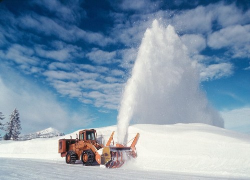 A snowplow clears a snowy road, sending a spray of snow into the air against a backdrop of blue sky and clouds.