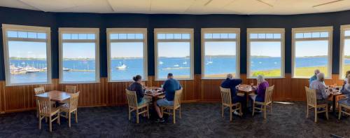 A restaurant with large windows overlooking a scenic waterfront, featuring diners enjoying their meals.