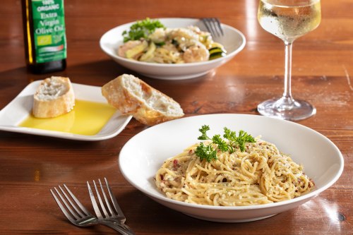 A bowl of creamy pasta with parsley, a glass of white wine, and a side of bread with olive oil on a wooden table.