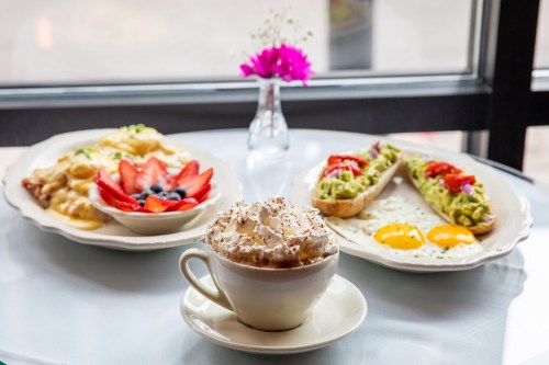 A table with a cup of whipped cream-topped coffee, a fruit dish, and two avocado toast plates with eggs and toppings.