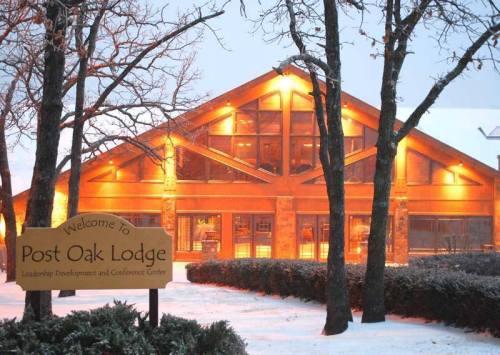 A warmly lit lodge with large windows, surrounded by snow-covered trees and a sign welcoming visitors to Post Oak Lodge.