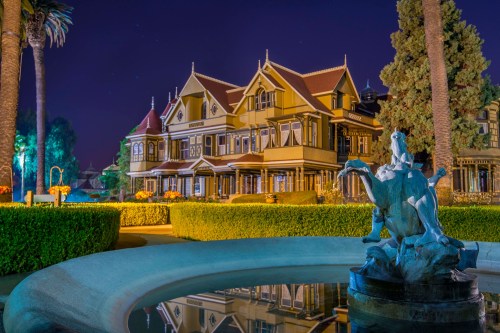The Winchester Mystery House at night, featuring a fountain and lush gardens in the foreground.