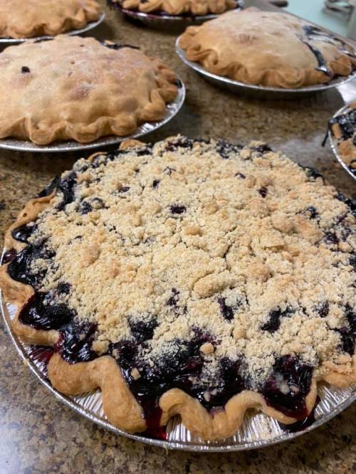 A freshly baked berry pie with a crumbly topping, placed on a countertop among other pies.