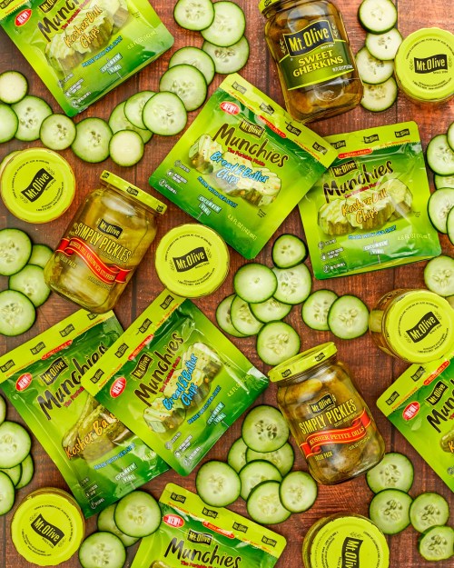 A flat lay of various pickles and cucumber slices on a wooden surface, featuring green packaging and jars.