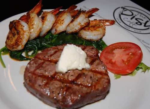 Grilled shrimp on spinach with a steak topped with butter and a slice of tomato on a white plate.